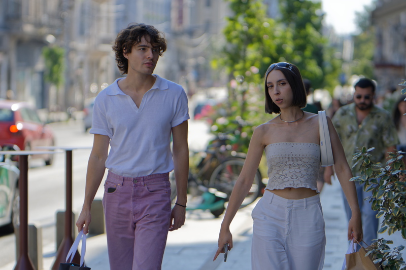 20 - Two individuals walking down a sunny city street, with the person on the left wearing a white collared shirt and pink trousers